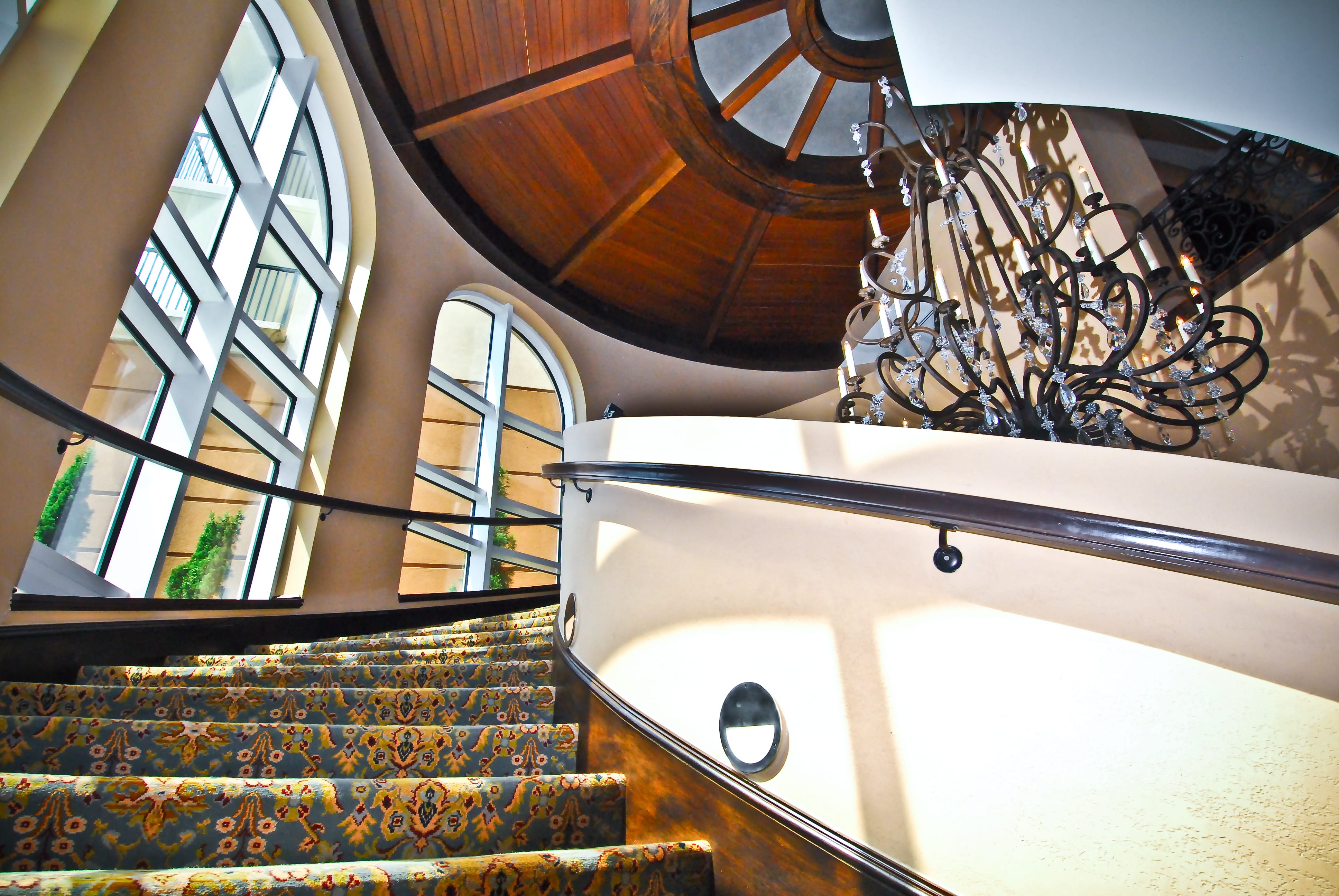 A curved staircase with carpeted steps and a chandelier in a lobby with arched windows.
