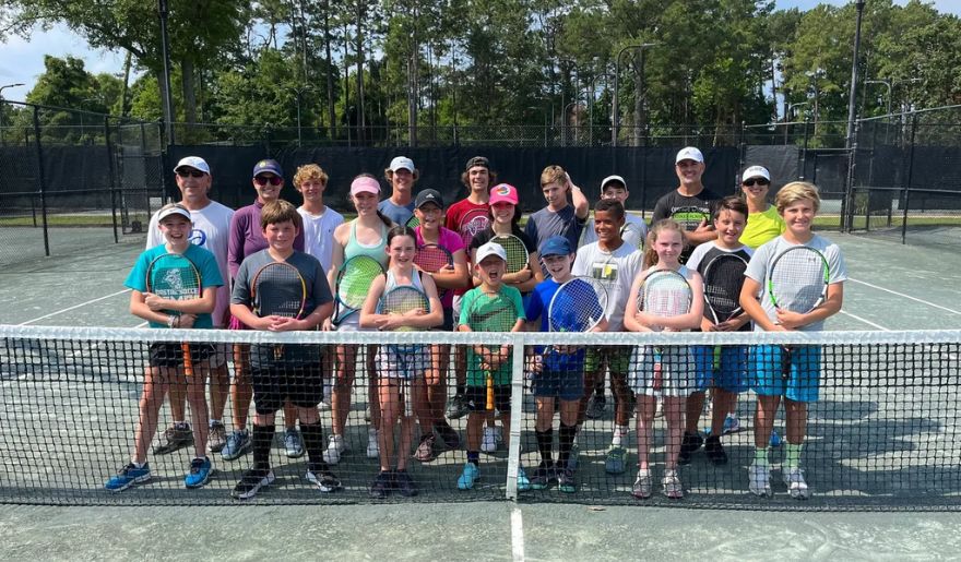 A joyful group of kids and adults pose for a picture on a tennis court at the Grande Dunes Tennis Club