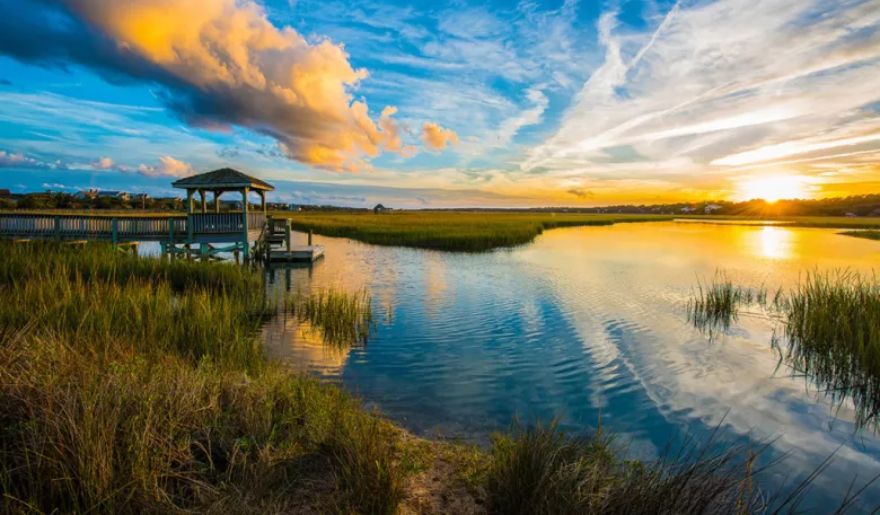 The sun sets beautifully over a tranquil marsh, highlighting a gazebo in the foreground at Huntington Beach State Park.