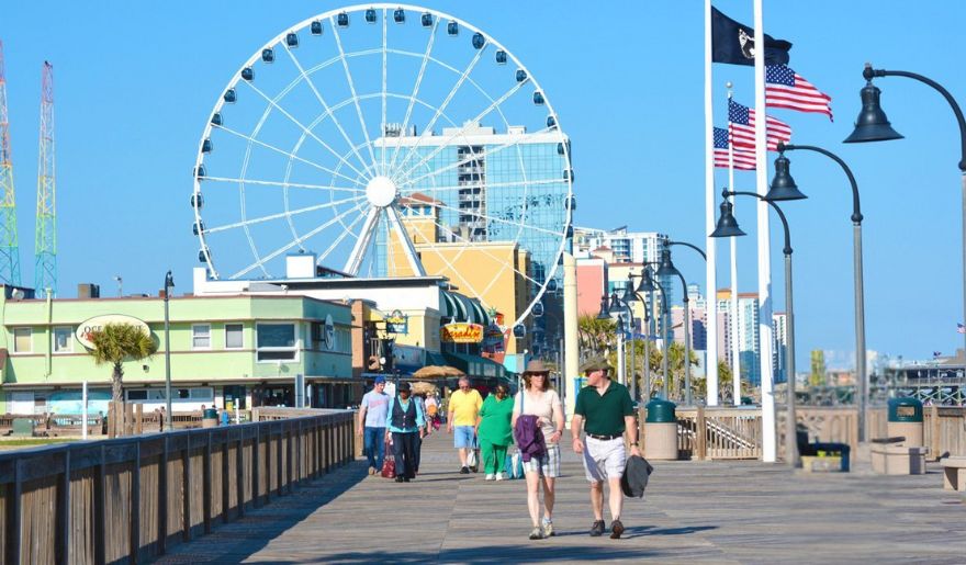 Couples stroll along the Myrtle Beach Boardwalk, with a vibrant Ferris wheel in the background, enjoying a scenic day out.