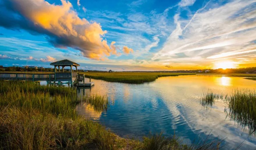 A serene sunset casts vibrant colors over the marsh and water at the end of the pier, creating a tranquil atmosphere.