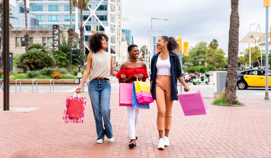 Three women walk along a street with shopping bags, enjoying a bachelorette party at Tanger Outlets in Myrtle Beach.