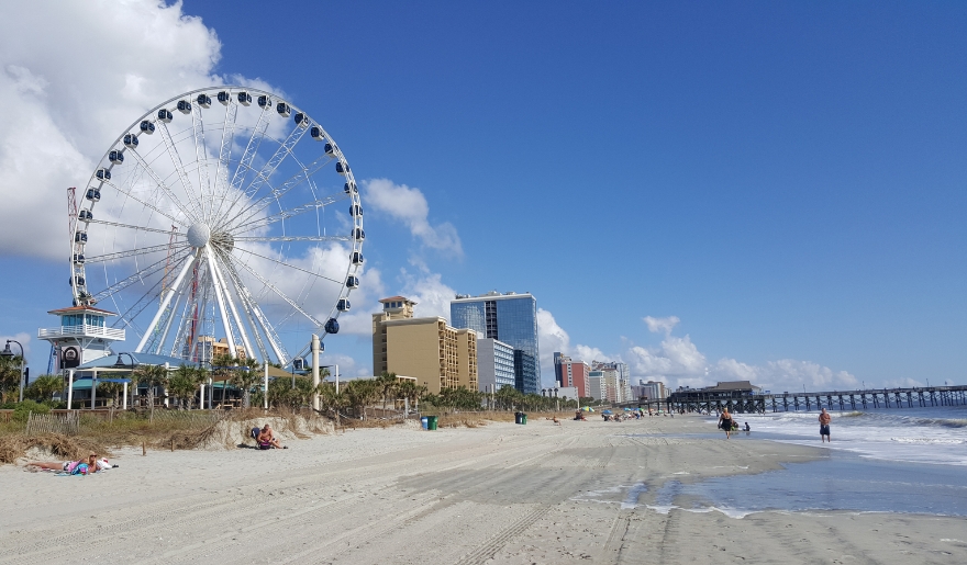 A vibrant ferris wheel stands on the beach, with families enjoying the sun and sand at Myrtle Beach's shoreline.