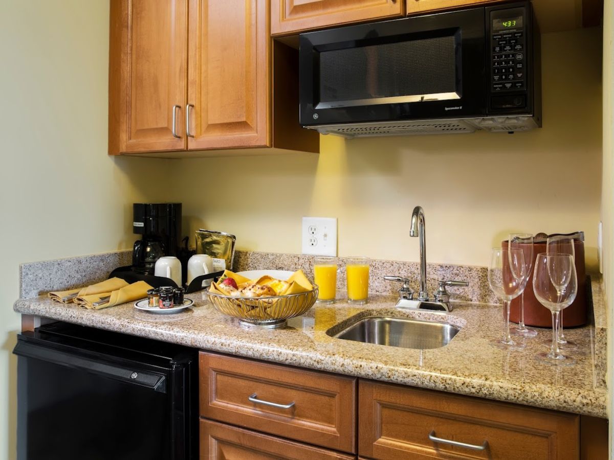 A kitchenette with a granite countertop, stainless steel sink, microwave, wine glasses, and a fruit bowl.