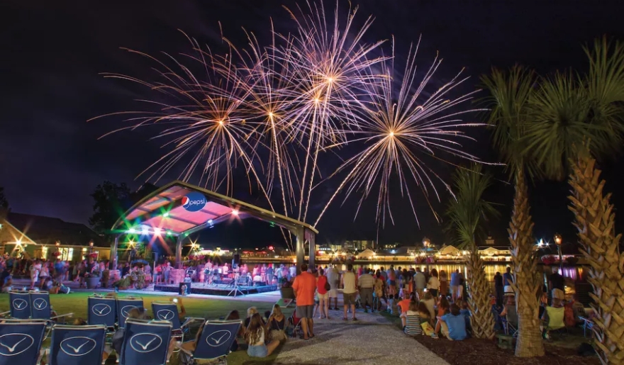 Colorful fireworks light up the night sky above the water, with attendees seated in lawn chairs at the Surfside Beach celebration.