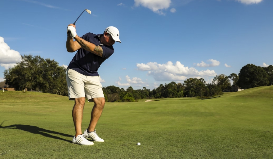 A golfer in a white hat and blue shirt swings his club, enjoying a sunny day on the golf course in Myrtle Beach.