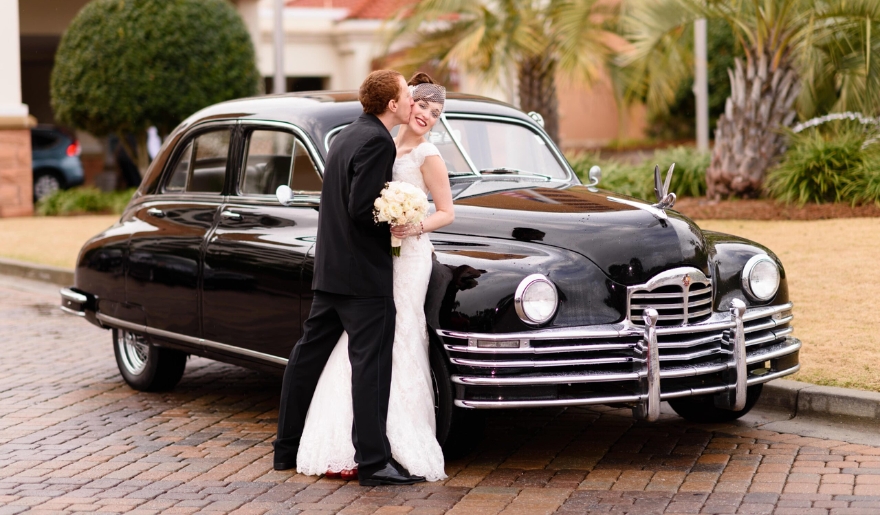 The bride and groom smile in front of a classic car, marking their special day at a Myrtle Beach wedding venue.