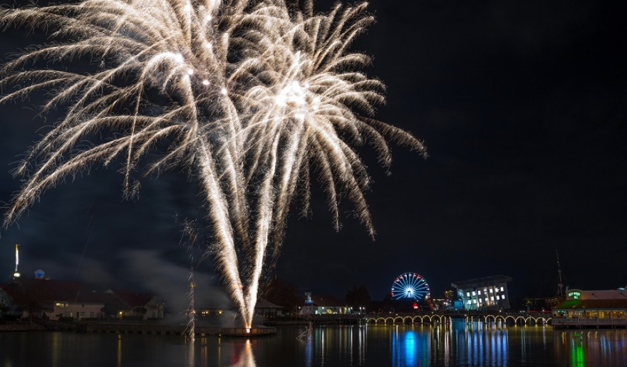 Vibrant fireworks burst over a lake, with a ferris wheel in view, during the August celebration at Broadway at the Beach.