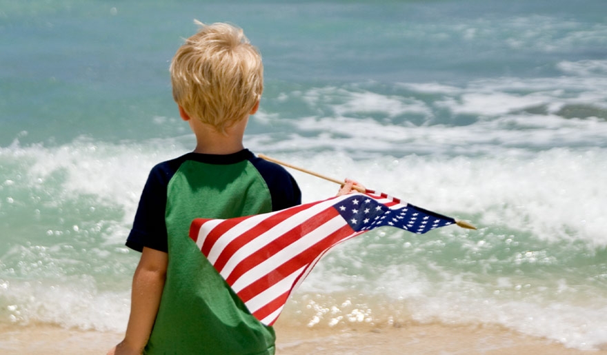 A kid holding an american flag, along the sandy beach, with gentle waves in the background.