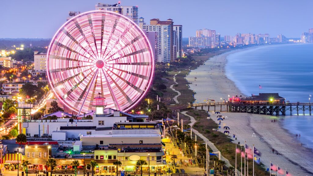 A vibrant Ferris wheel lights up the Myrtle Beach boardwalk at dusk, with the beach and ocean extending into the distance.
