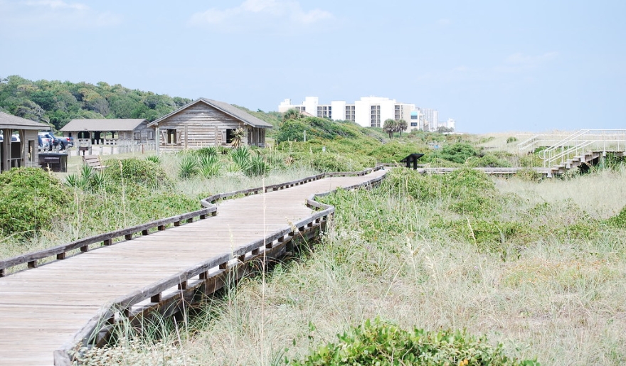 A wooden boardwalk leads to a sandy beach with a house in the background, highlighting Myrtle Beach's outdoor charm.