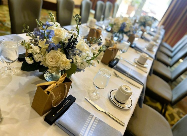 A long formal dining table set with white and blue floral centerpieces and gift bags.
