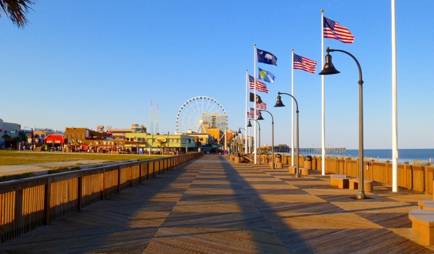 View along the Myrtle Beach Boardwalk showing oceanfront walkway, American flags, nearby shops, and the SkyWheel in the distance, representing popular free attractions.