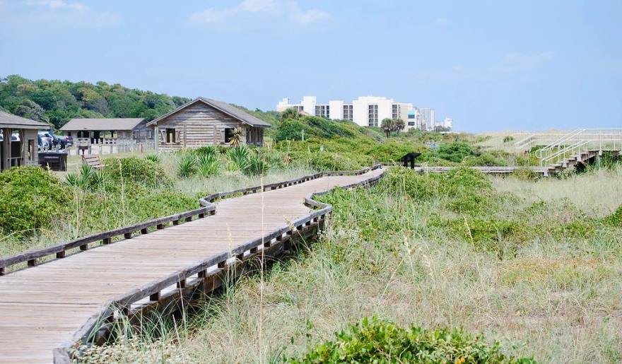 A wooden boardwalk extends to a beach, with a house visible nearby, highlighting family activities at Myrtle Beach State Park.