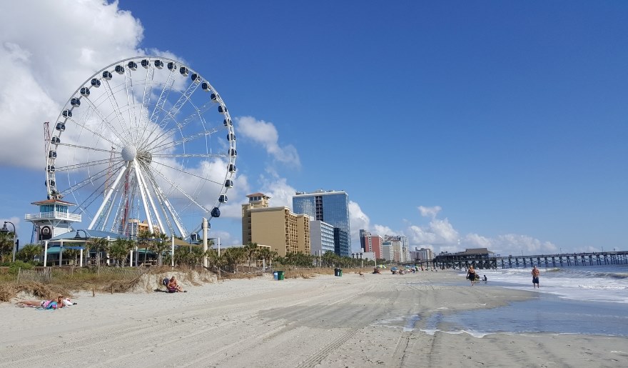 A vibrant ferris wheel stands on the beach, with families enjoying the sun and sand at Myrtle Beach's shoreline.