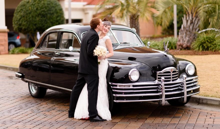 The bride and groom smile in front of a classic car, marking their special day at a Myrtle Beach wedding venue.