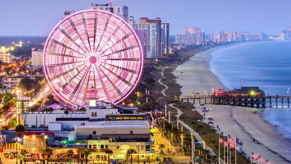 A vibrant Ferris wheel lights up the Myrtle Beach boardwalk at dusk, with the beach and ocean extending into the distance.