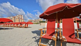 Red umbrellas and lounge chairs lined up on a sandy beach in front of coastal resort buildings.