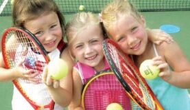 Three smiling young girls holding tennis rackets and balls on a tennis court.