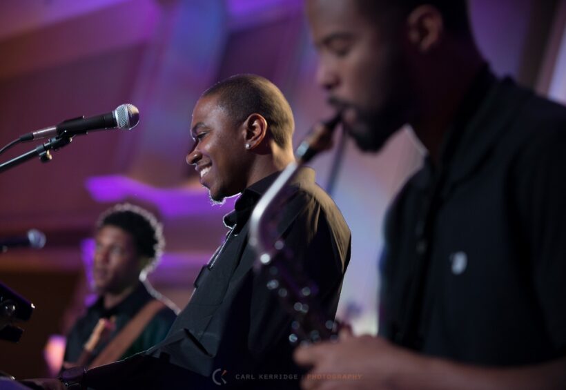 Musicians performing live on stage, including a smiling keyboardist and a saxophonist.