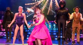 A lead singer in a bright pink dress performing on stage with costumed backup dancers.