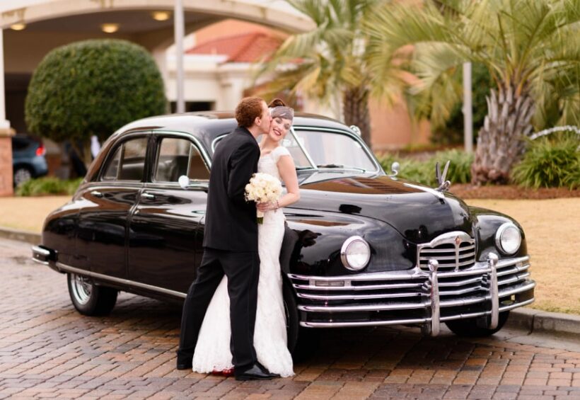 A groom kissing his bride on the cheek next to a classic black vintage car.
