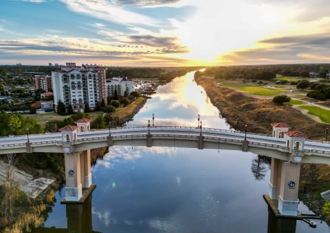 Aerial view of a bridge arching over a calm river at sunset, next to waterfront buildings.